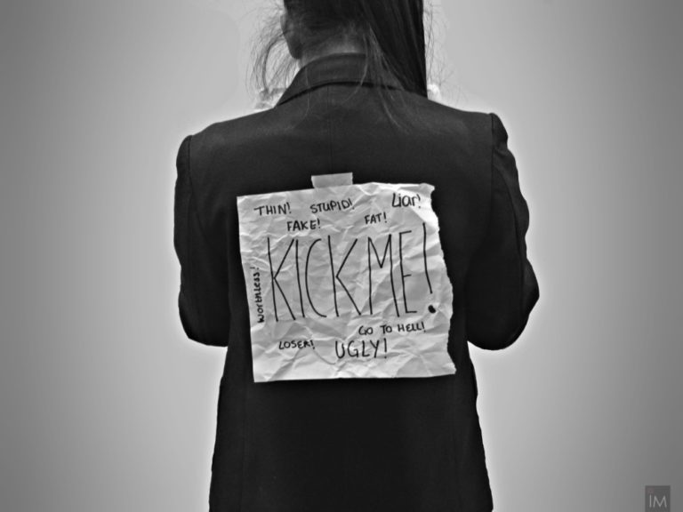 Foto preta e branca com uma menina com um cartaz nas costas escrito "me chute".